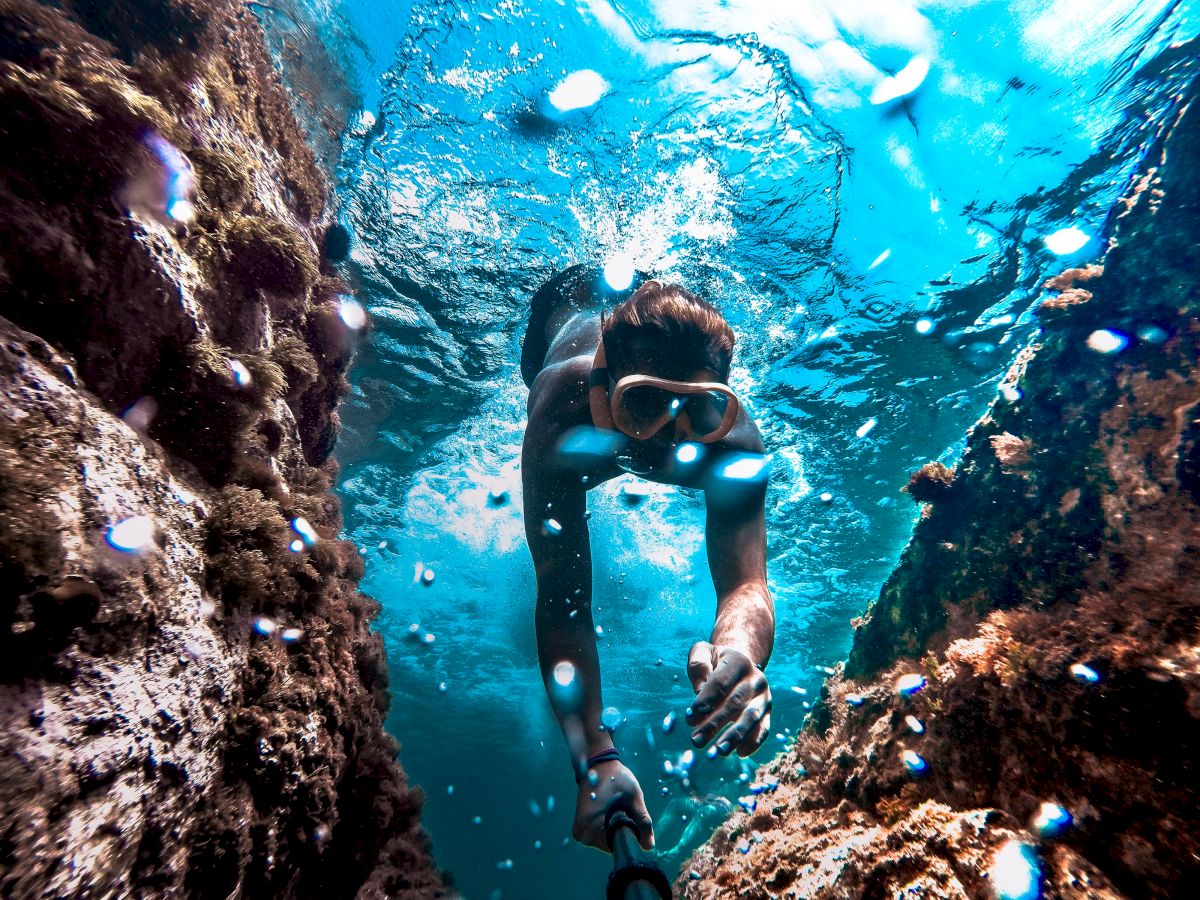 A snorkeler swims underwater between rocks, wearing a mask and snorkel, with sunlight filtering through the clear blue water.