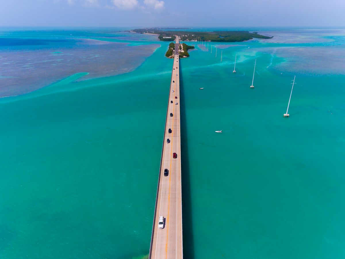 Aerial view of a long, narrow bridge extending over turquoise water, connecting small land masses. The bridge has vehicles traveling on it.