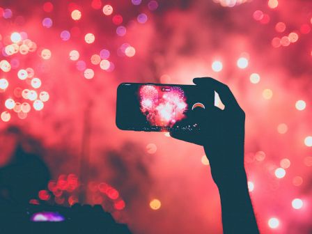 A hand holding a smartphone captures colorful fireworks in the sky, with vibrant red and pink lights creating a dazzling display.