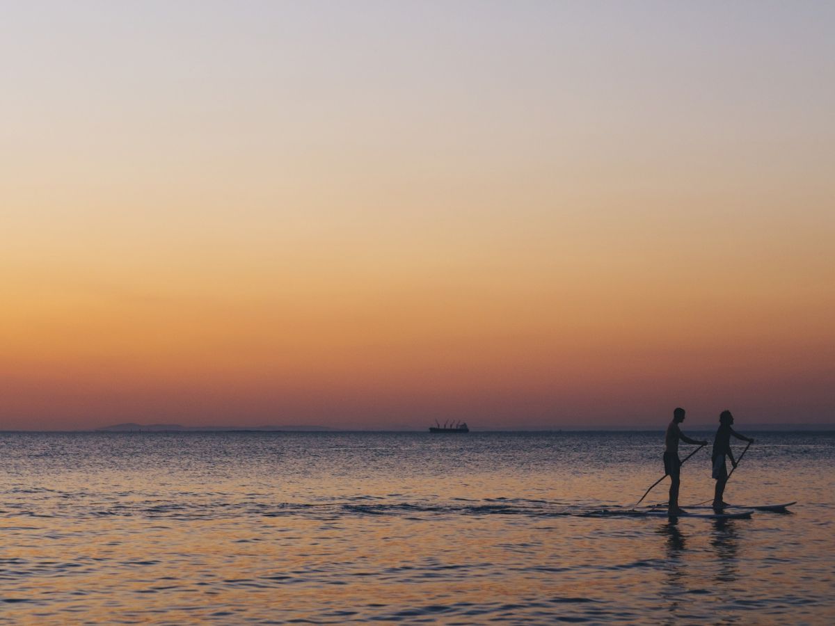 Two people paddleboarding on calm water during sunset, with an orange to purple gradient sky and a distant boat on the horizon.