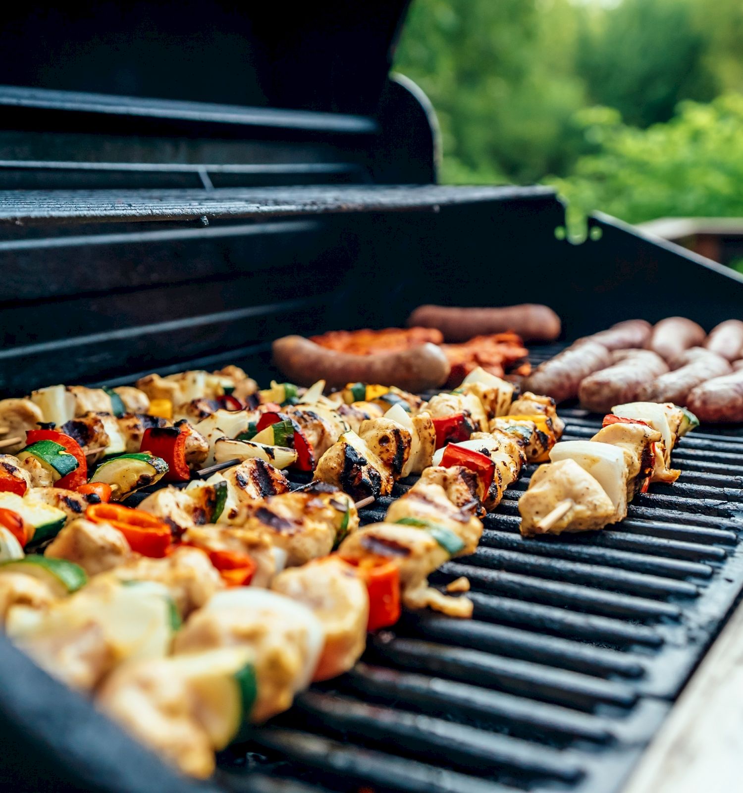 A grill loaded with kebabs and sausages, set against a blurred background of greenery, capturing a summer barbecue scene.