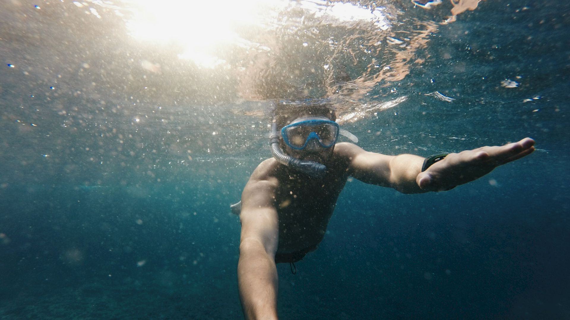 A person is snorkeling underwater, wearing a mask and snorkel, reaching out one arm as sunlight filters through the surface of the water.
