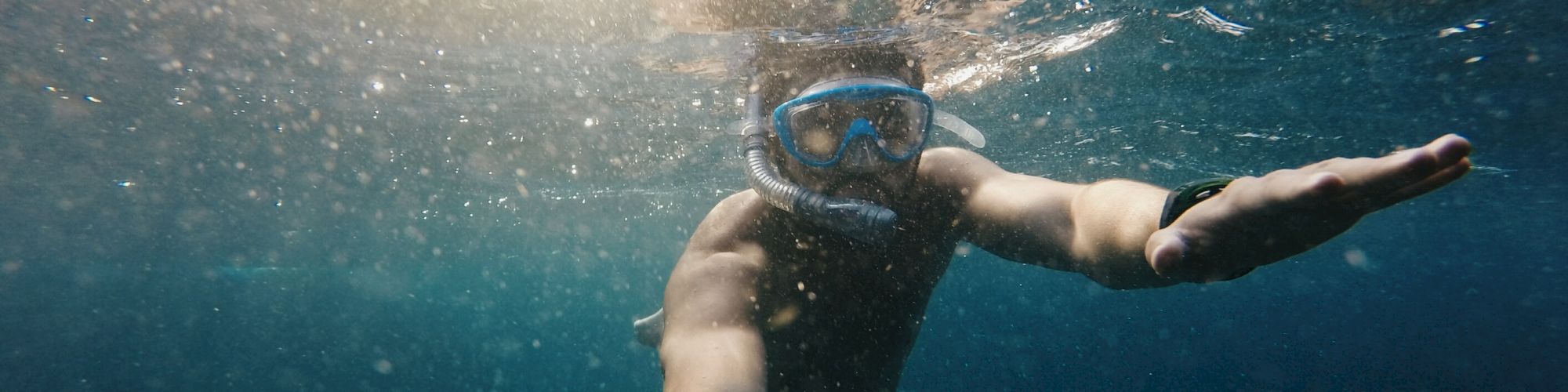 A person is snorkeling underwater, wearing a mask and snorkel, reaching out one arm as sunlight filters through the surface of the water.