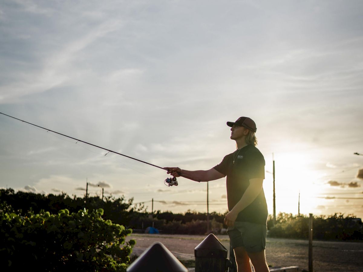 A person is fishing from a dock at sunset, casting a fishing rod with a serene backdrop of a glowing sky and silhouetted trees.