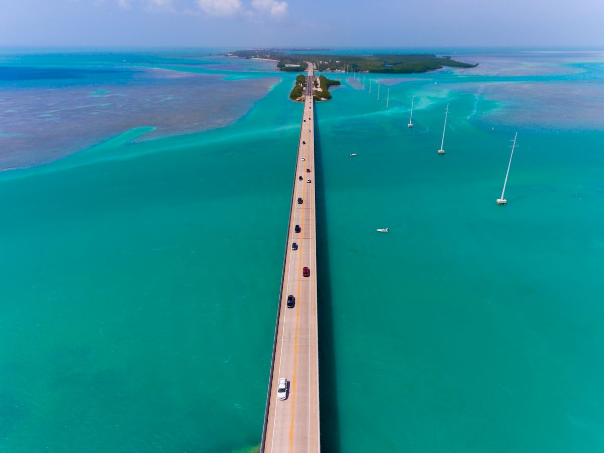 A long, straight bridge over turquoise waters, connecting islands with cars driving on it under a clear blue sky.