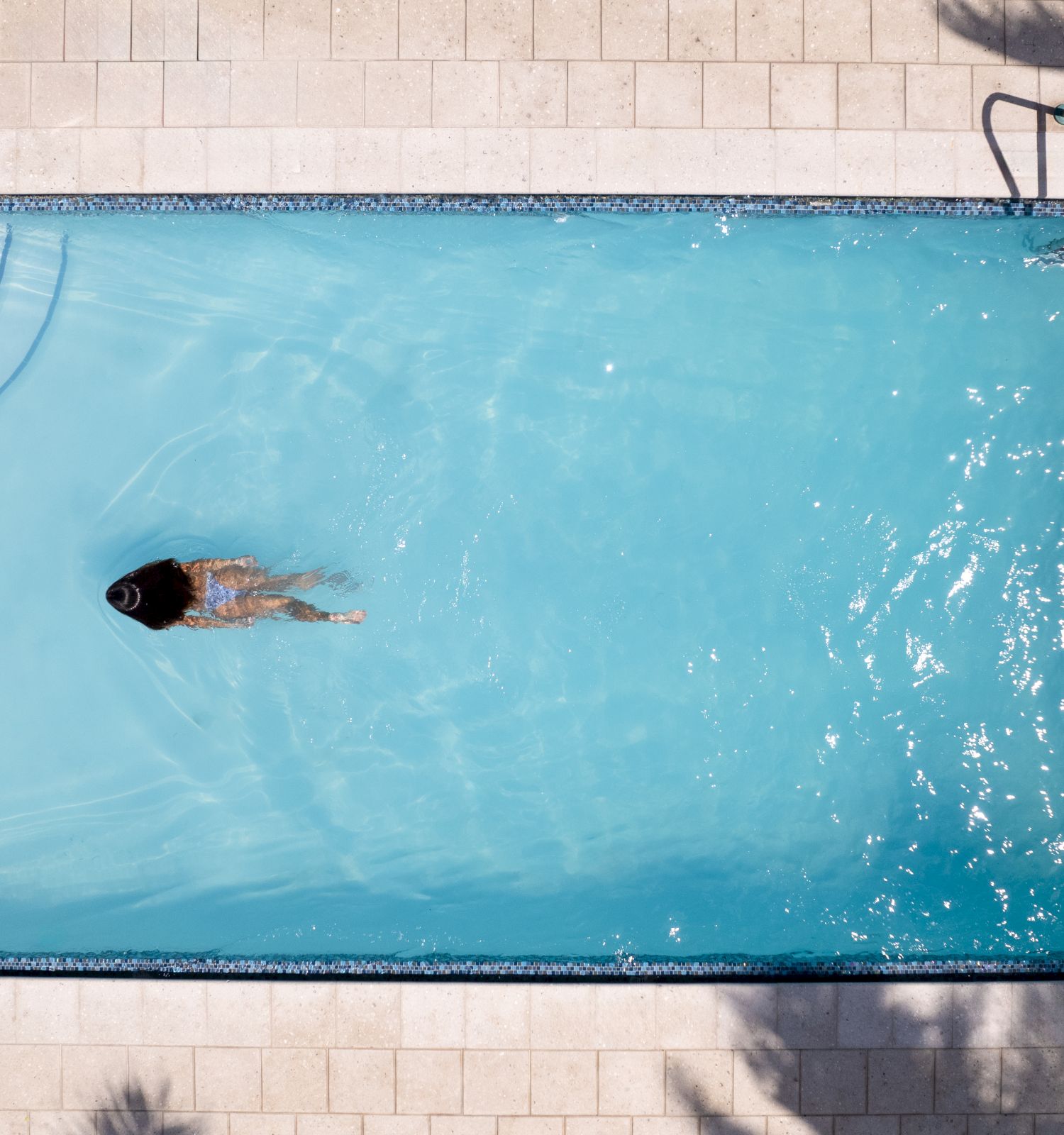 A person is swimming in a rectangular pool, viewed from above. The pool has a ladder and steps in one corner, with palm shadows visible.