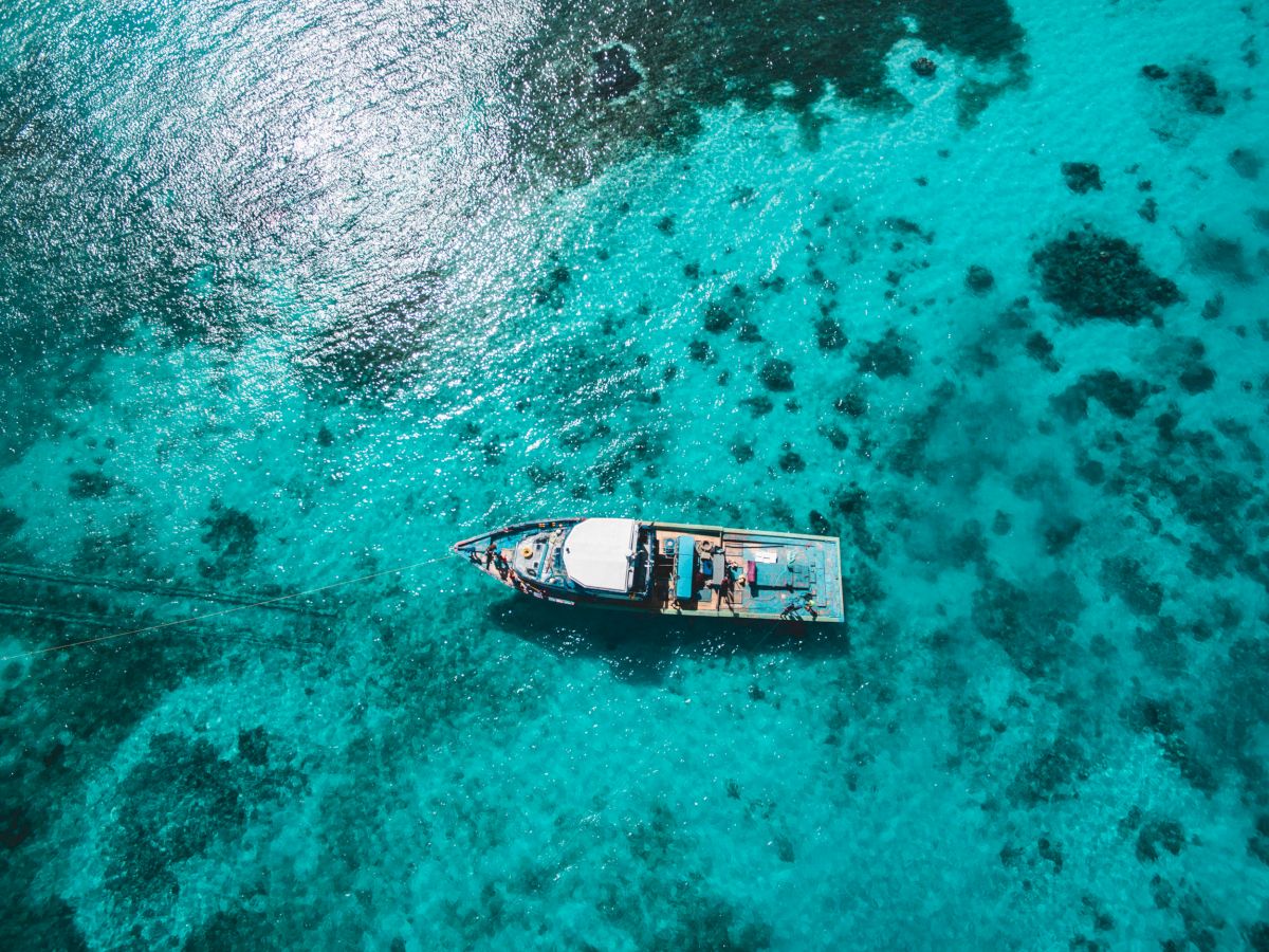 An aerial view of a boat floating on clear turquoise water, surrounded by visible patches of coral reefs and marine life.