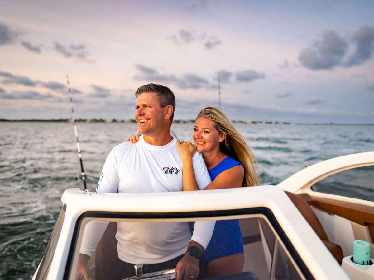 A man and woman smile while on a boat with an ocean backdrop during sunset, with fishing poles visible in the background.