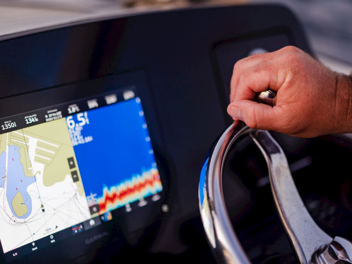 A hand is gripping a boat's steering wheel, and a GPS/navigation system screen shows maps and data.