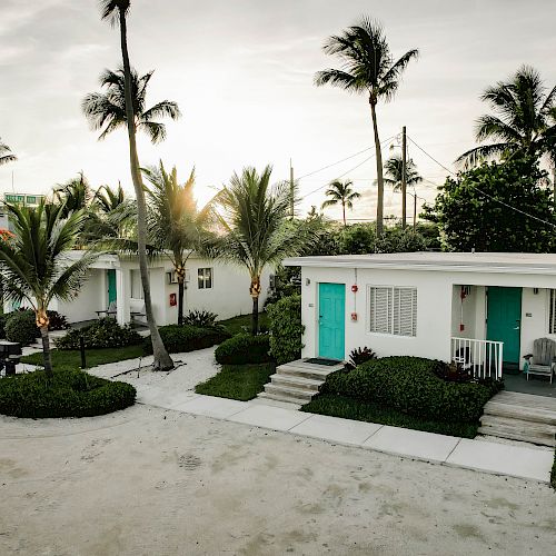 The image shows a serene scene with small, white bungalows with turquoise doors, surrounded by palm trees and greenery, possibly in a tropical location.