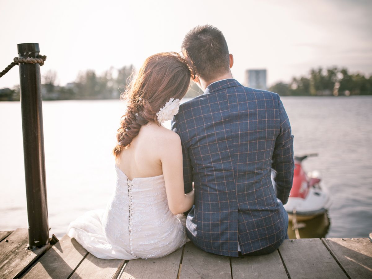 A couple in formal attire sits closely on a wooden dock, overlooking a calm body of water, with a jet ski in the background at sunset.