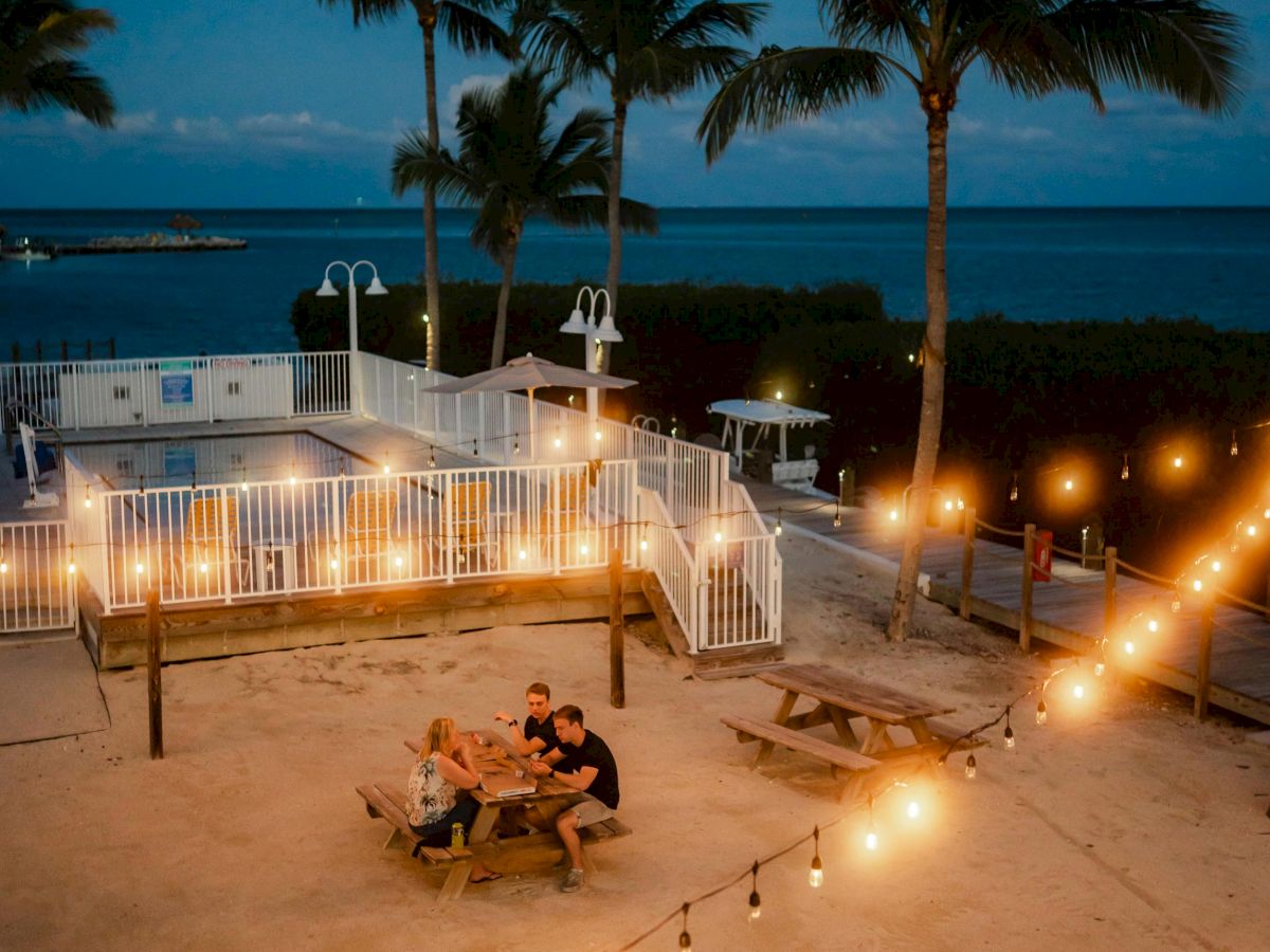 An evening seaside setting with string lights, palm trees, and people sitting at picnic tables, creating a cozy and relaxed beachside atmosphere.