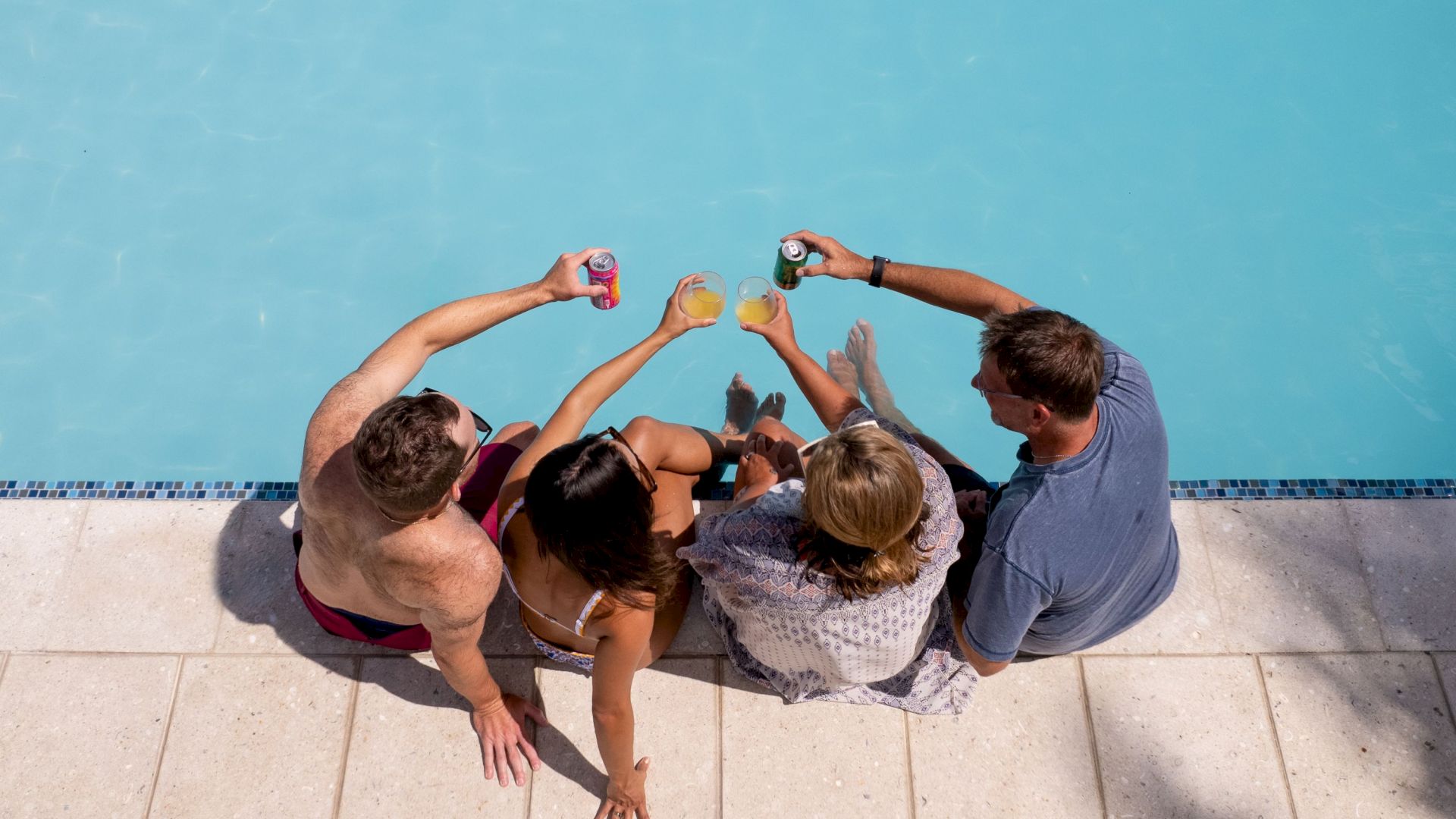 Four people are sitting by a pool, raising their drinks in a toast, viewed from above.