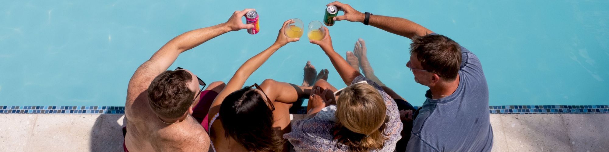 Four people are sitting by a pool, raising their drinks in a toast, viewed from above.