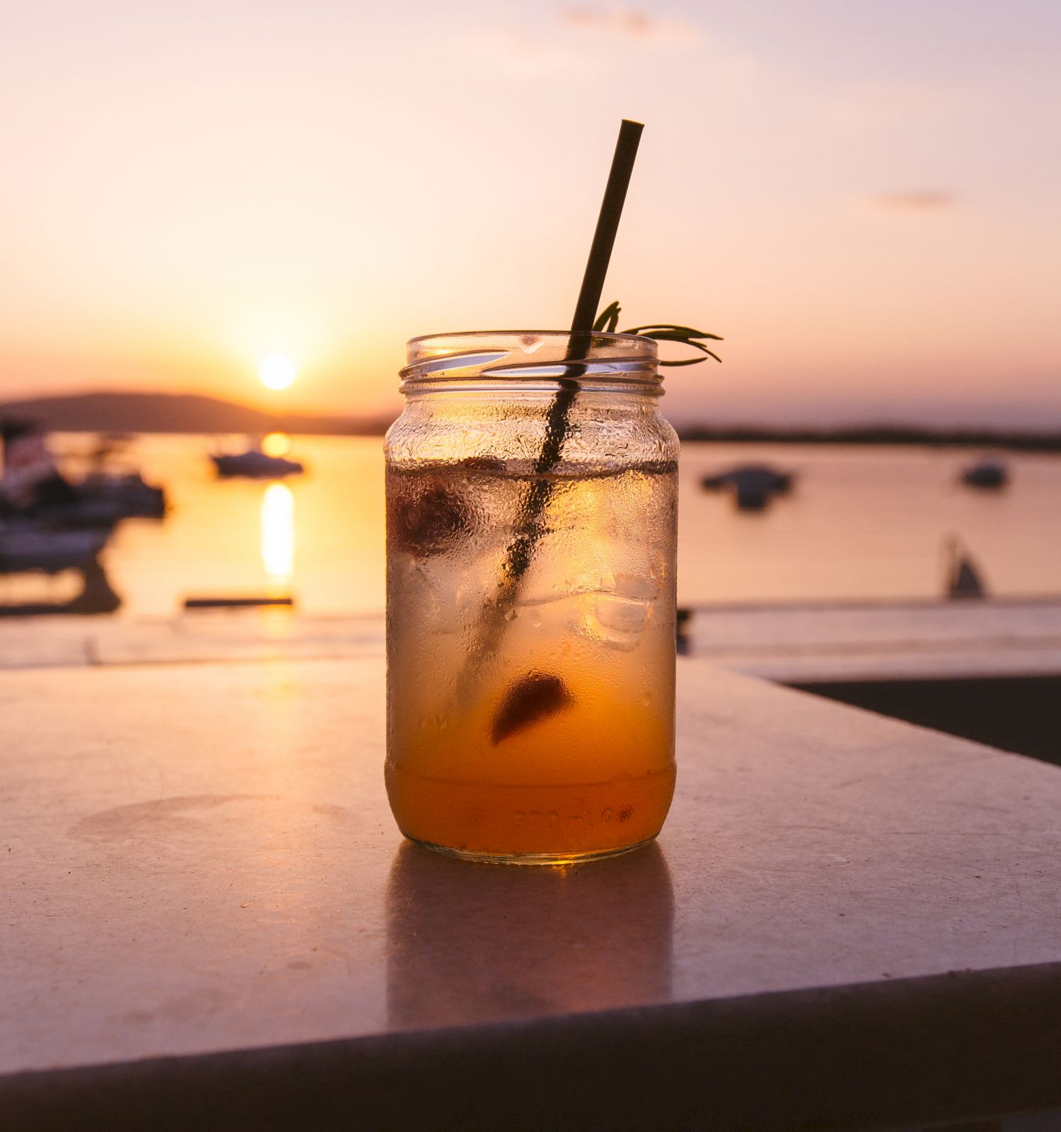 A mason jar with a drink and a straw sits on a table, with a sunset and boats in the background.