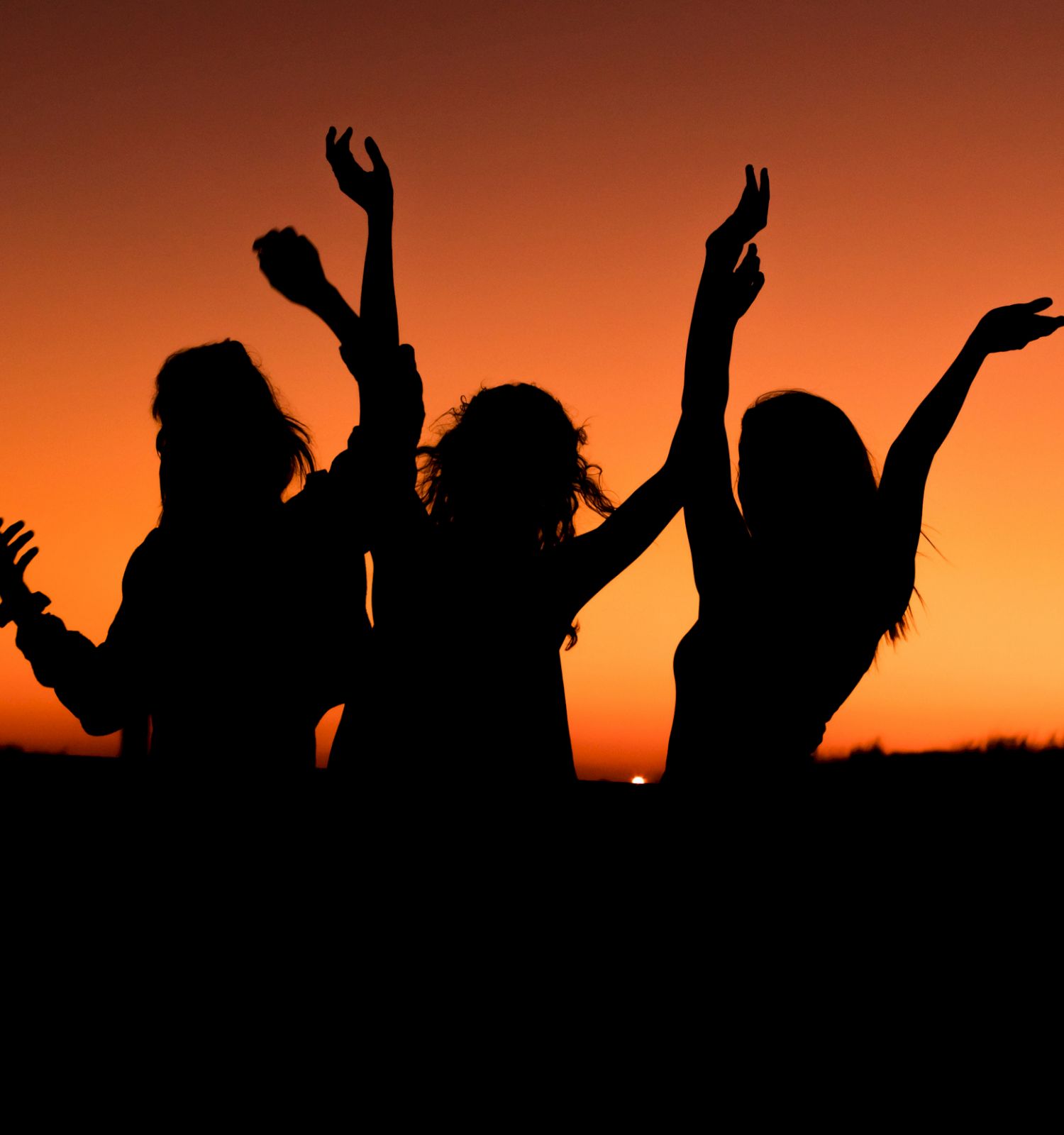 Three silhouettes of people with arms raised are set against a vibrant orange sunset sky, creating a joyous and carefree scene.