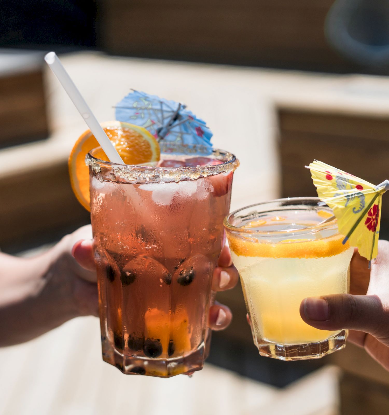 Two people are holding colorful cocktails with straws, fruit garnishes, and decorative umbrellas, seemingly toasting.