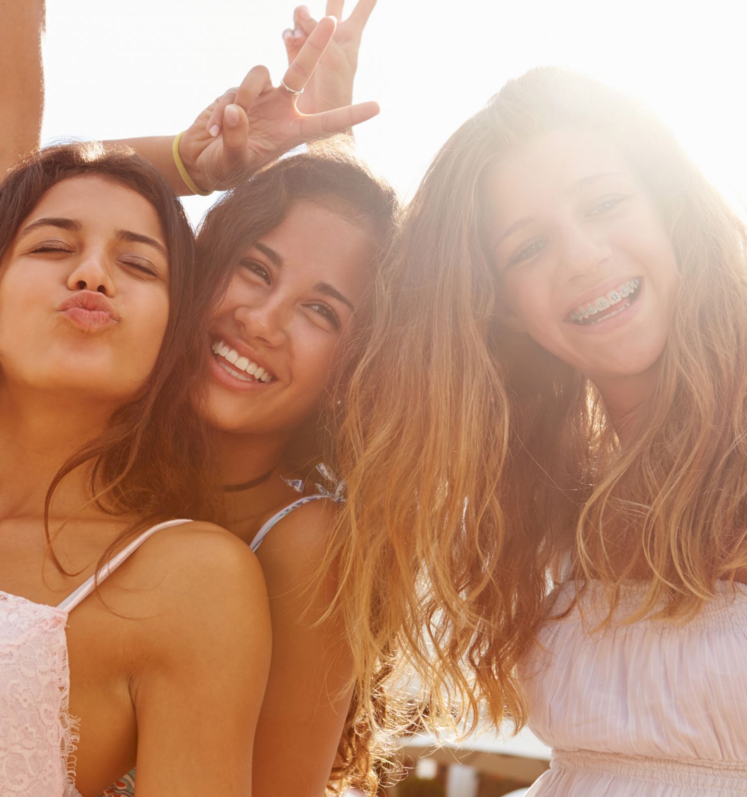 Three people are posing for a selfie outdoors, smiling and making playful gestures, with bright sunlight in the background.