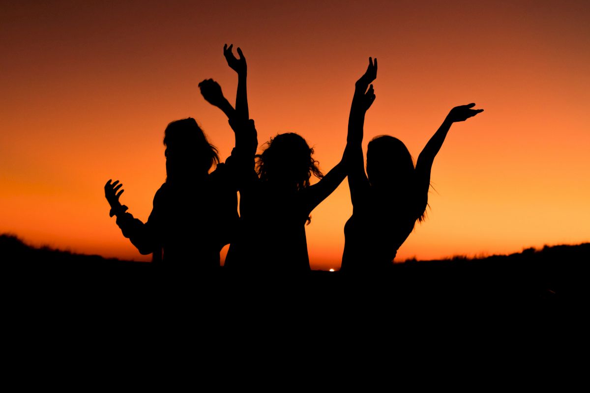 The image shows three silhouetted figures with outstretched arms against an orange sunset sky, creating a lively and celebratory scene.