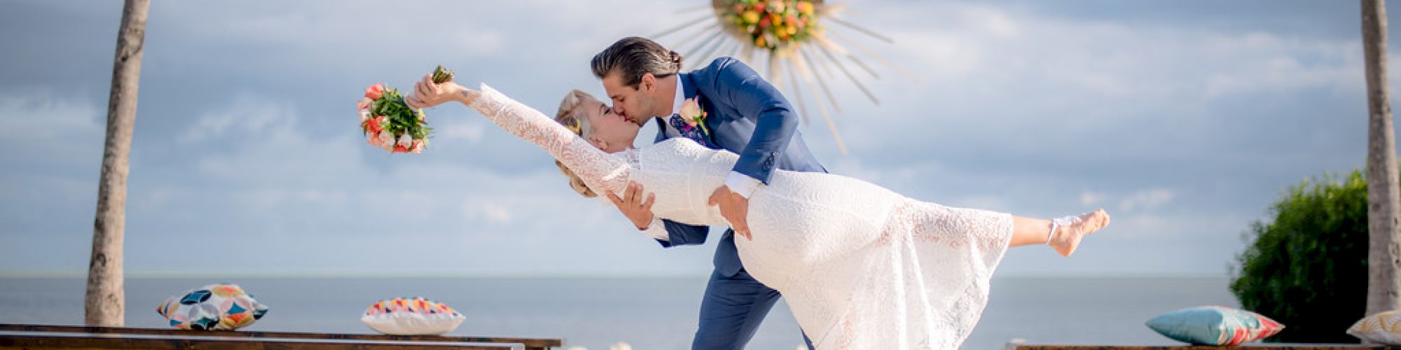 A couple dressed in wedding attire is posing on a beach, with the groom dipping the bride. Palm trees and benches are in the background.