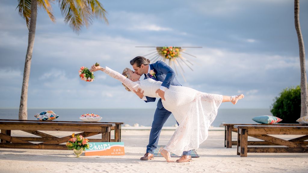 A couple dressed in wedding attire is posing on a beach, with the groom dipping the bride. Palm trees and benches are in the background.