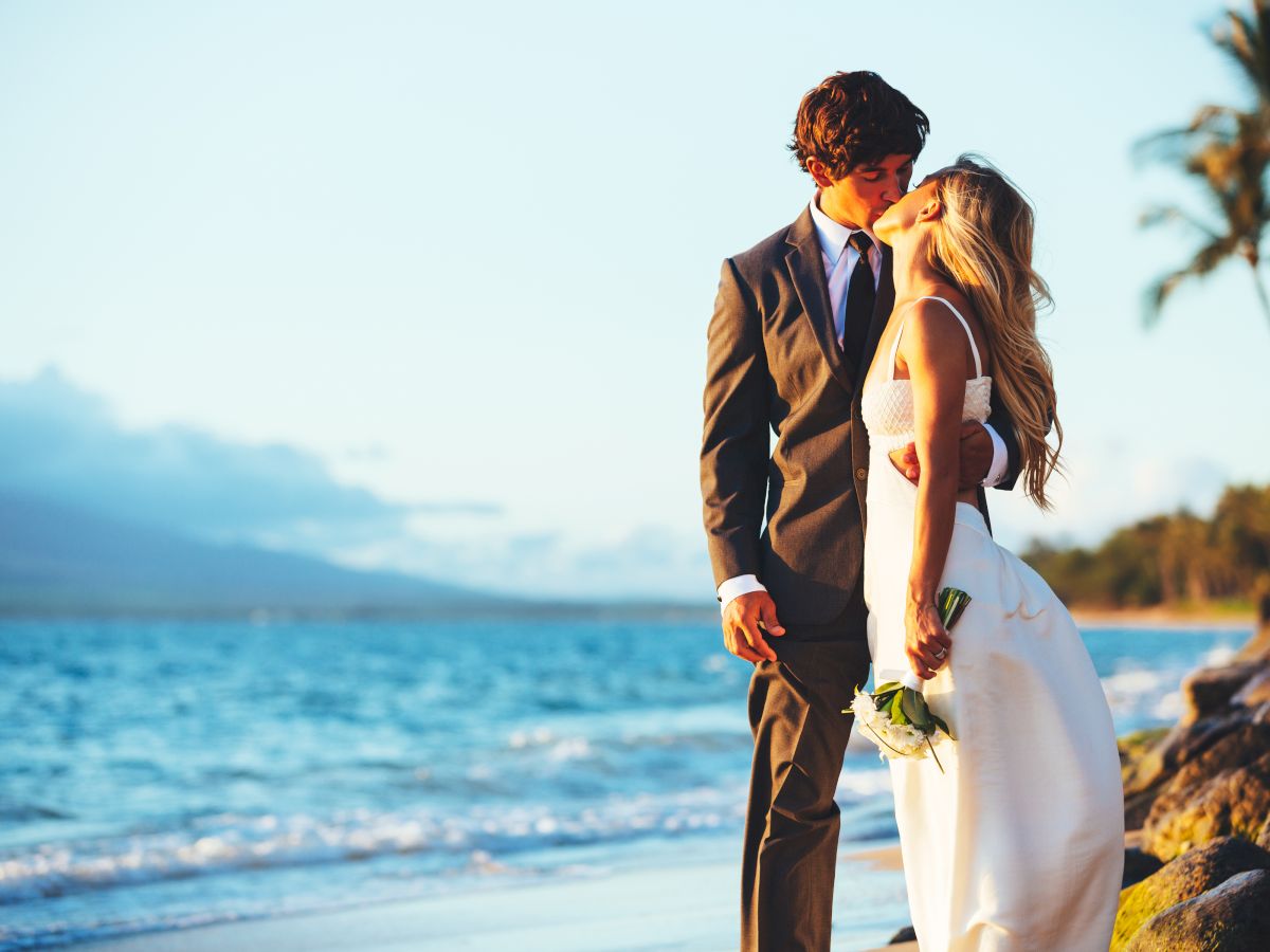 A couple in wedding attire is kissing on a beach with the ocean and palm trees in the background during sunset.