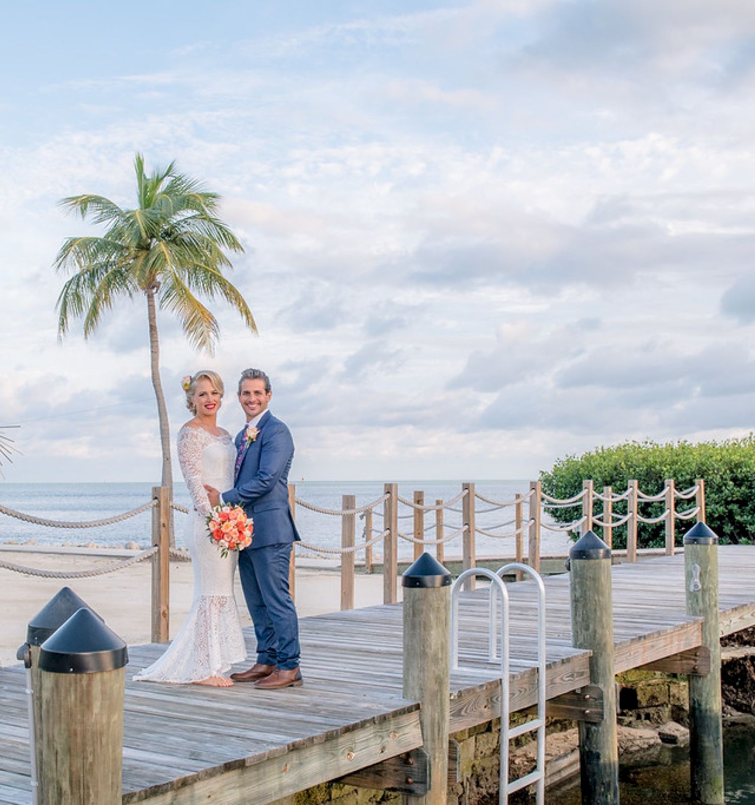 A couple in wedding attire stands on a wooden dock by the water, with palm trees and a tropical beach in the background.