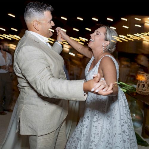 A couple is joyfully dancing at a lively event with string lights overhead, surrounded by people and festive decorations.