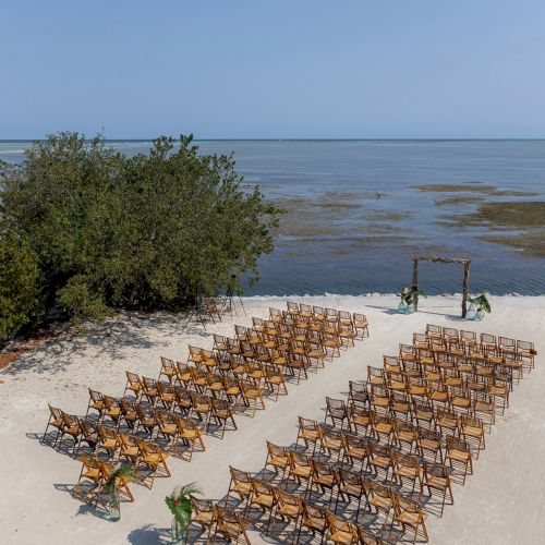 Outdoor wedding setup with wooden chairs arranged on a sandy beach facing an altar, near the sea and some greenery.