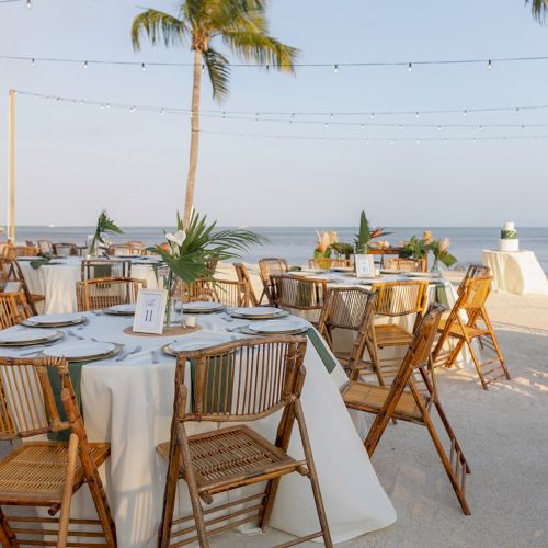 An outdoor event setup with round tables, wooden chairs, and palm trees, overlooking the ocean under string lights.