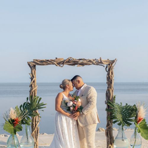 A couple stands under a rustic wooden arch on the beach, framed by tropical plants, against a serene ocean backdrop, sharing a tender moment.