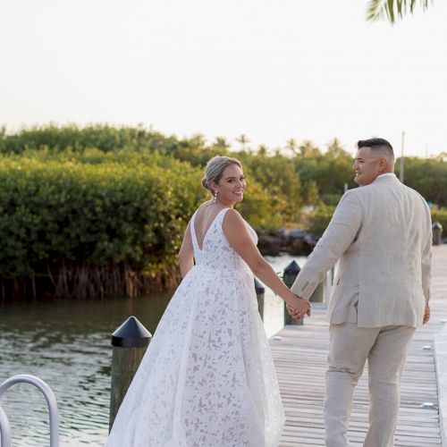 A couple in wedding attire is walking on a wooden dock near the water, surrounded by greenery, holding hands.