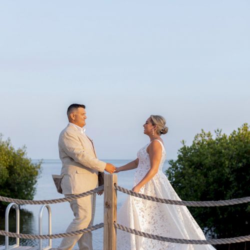 A couple in wedding attire stands on a wooden dock, holding hands with a scenic water backdrop and greenery.