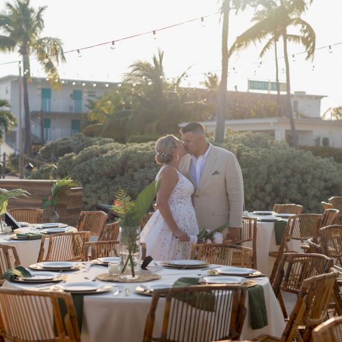 A couple stands in a sunlit outdoor setting, surrounded by decorated tables and chairs, with palm trees in the background.