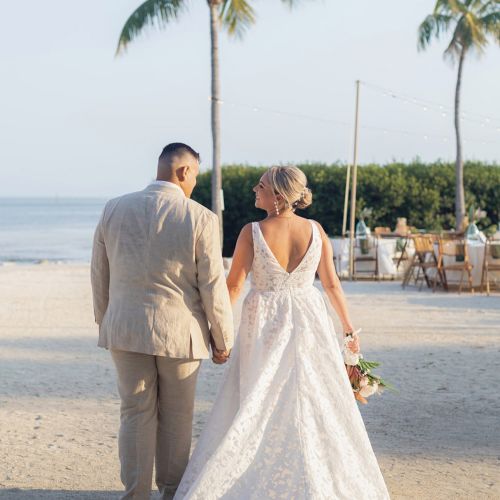 A couple in wedding attire holding hands on a beach, surrounded by palm trees, with a view of the ocean.