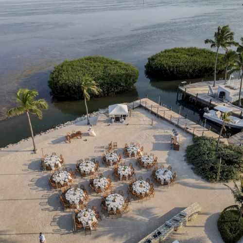 An outdoor event setup on a beach with tables and chairs, near a pool and palm trees by the water, creating a scenic atmosphere.