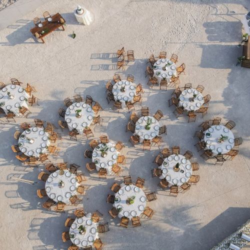 The image shows an outdoor event setup with round tables and chairs arranged on a sandy surface, possibly for a wedding or gathering.