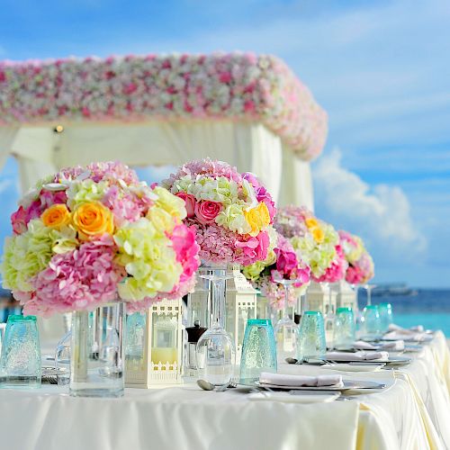 An elegant beachside table setup with vibrant floral centerpieces, adorned with pastel drapery, overlooking the ocean on a clear day.