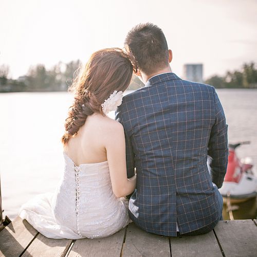 A couple sits on a wooden pier by the water, facing away, in formal attire. The serene and romantic setting includes a jet ski in the background.