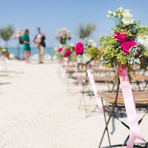 Outdoor wedding setup with chairs decorated with floral arrangements and ribbons, two people standing in the background near the altar.