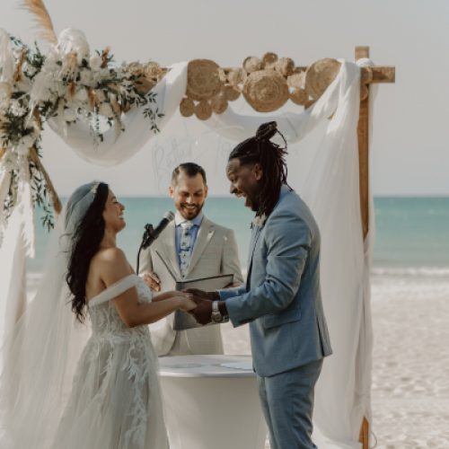 A couple exchanges rings during their wedding ceremony on a beach, with an officiant standing behind them under a decorated canopy, facing the ocean.