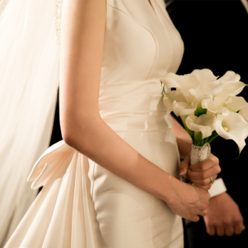 A bride in a white dress is holding a bouquet of white flowers, standing next to a person in a black suit, likely during a wedding ceremony.