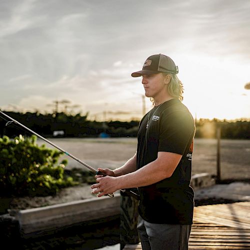 A person in a hat and t-shirt is fishing by a dock during sunset, with greenery and a partly cloudy sky in the background.