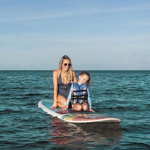 A woman and a child are sitting on a paddleboard in calm ocean water, both wearing swimwear. The sky is clear with light clouds.