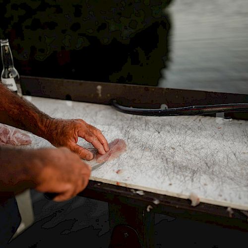 A person is filleting a fish on a table near the water, with a knife and a drink bottle beside them.