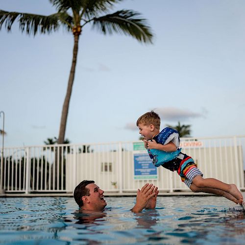 A man in a swimming pool is about to catch a boy who is joyfully jumping toward him. Palm trees and a fence are visible in the background.