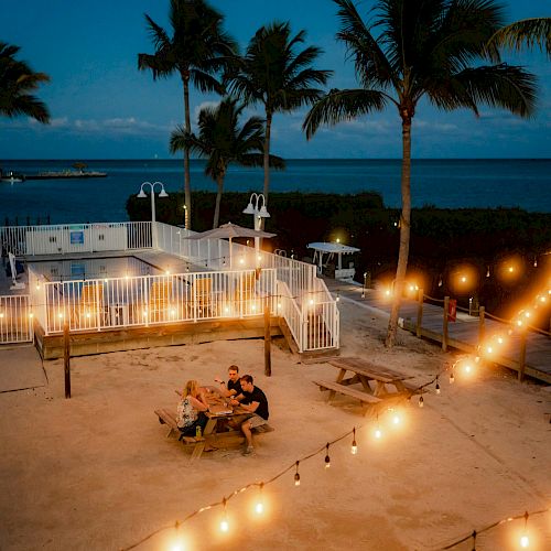 Outdoor dining scene with string lights, palm trees, and ocean view at dusk. People seated at picnic tables enjoying dinner. Peaceful atmosphere.