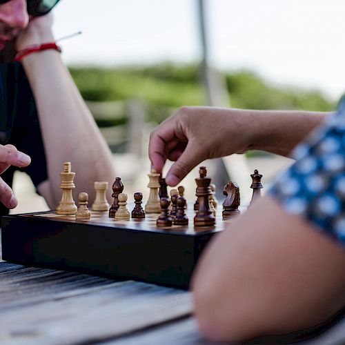 Two people are engaged in a game of chess on an outdoor wooden table, with one person making a move.