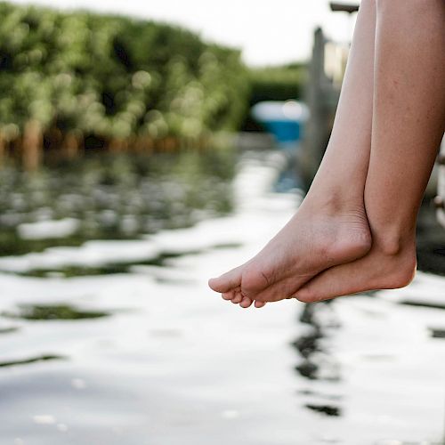 A pair of feet dangle from a wooden dock above a calm body of water with greenery in the background.