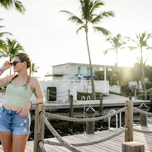 A woman in sunglasses stands on a wooden dock, surrounded by palm trees and buildings, enjoying a sunny day by the water.